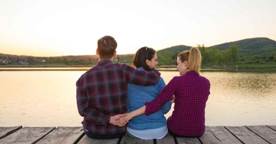 A man and two women sitting on a pier. The man has his arm around one woman, presumably his wife, but his other hand is holding the second woman's hand behind his wife's back