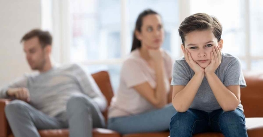A young boy with his head in his hands. His parents sit in the background facing away from each other