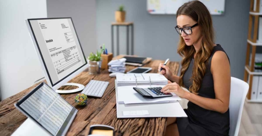 A woman sitting at a desk looking at a computer. She is wearing glasses and doing calculations in a notebook with a calculator