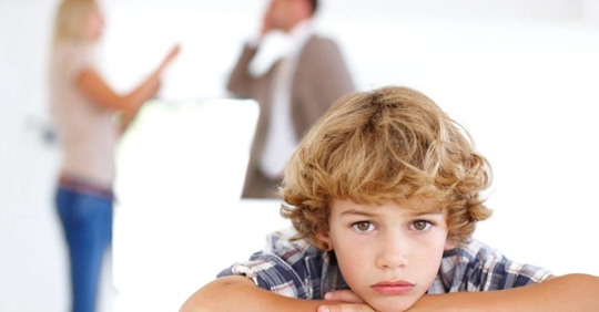 A child sadly resting his head on his hands while his parents argue in the background.