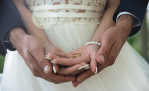 A groom stands behind his bride with his arms around her. Their hands are stacked and she is holding two gold wedding bands.